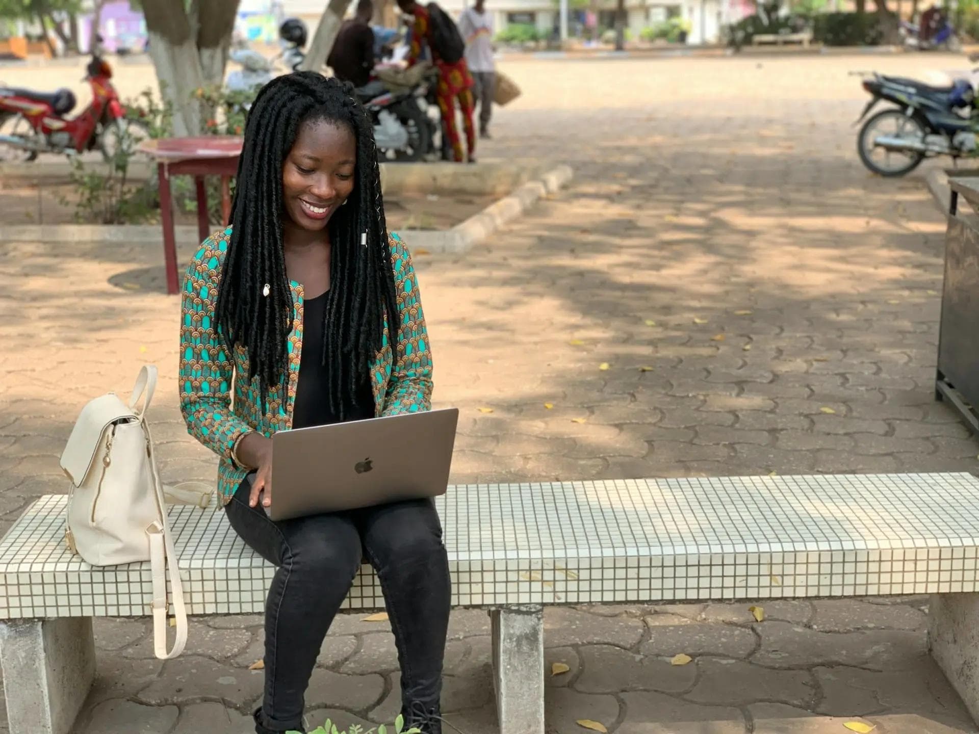 Black woman with braids outside on bench in park typing on laptop